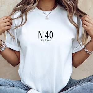 May include: White t-shirt with the text "N&deg;40 ORIGINAL EST 1986" in black. The shirt is a classic crew neck style. The model is wearing blue jeans and white sneakers.