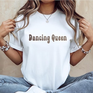 May include: A white t-shirt featuring the text "Dancing Queen" in a brown, glittery font. The shirt is paired with blue jeans and white trainers. The person is wearing bracelets and a necklace.