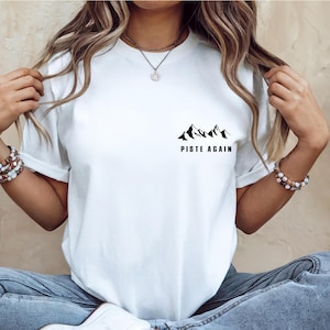 May include: White t-shirt with a black mountain graphic and the text "PISTE AGAIN". The shirt is made of a soft material and is suitable for casual wear.