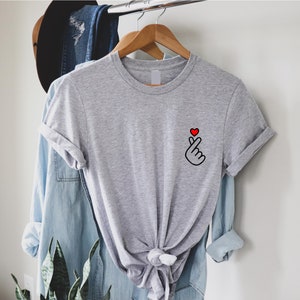 May include: A grey t-shirt with a small, black and red hand gesture graphic on the left chest. The hand is making a heart shape with the index and pinky fingers, and the thumb is pointing up. The heart is red.