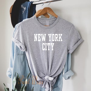 Puede incluir: Camiseta gris con texto blanco que dice "NEW YORK CITY". La camiseta est&aacute; colgada en una percha de madera.