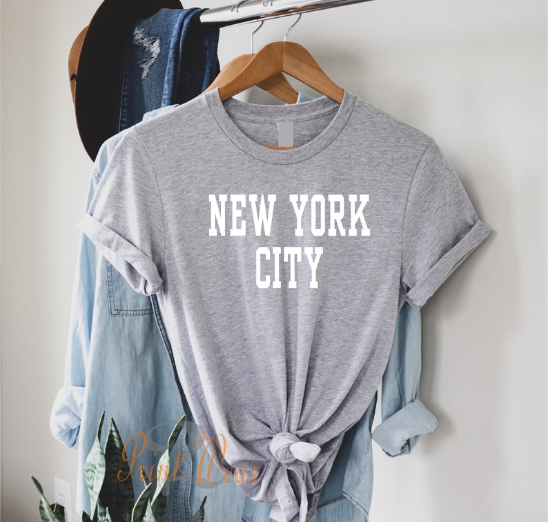 Puede incluir: Camiseta gris con texto blanco que dice "NEW YORK CITY". La camiseta est&aacute; colgada en una percha de madera.