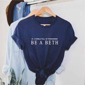 Puede incluir: Una camiseta azul marino con texto blanco que dice "En un mundo lleno de princesas, sé una Beth."