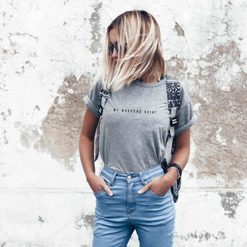 May include: A woman wearing a gray t-shirt with the text "My Weekend Shirt" and blue jeans. She is standing in front of a white wall.