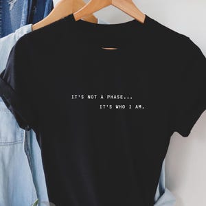 May include: A black short-sleeved t-shirt featuring white text printed on the chest that reads, IT'S NOT A PHASE... IT'S WHO I AM. The garment has a crew neckline and is displayed on a wooden hanger, with a light blue denim fabric visible behind it.