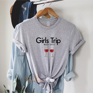 Pode incluir: Camiseta cinza com o texto "Girls Trip" e "Ibiza 2024" impresso nela. Duas ta&ccedil;as de vinho tinto com glitter est&atilde;o abaixo do texto.
