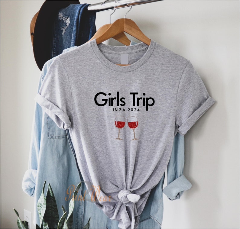 Pode incluir: Camiseta cinza com o texto "Girls Trip" e "Ibiza 2024" impresso nela. Duas ta&ccedil;as de vinho tinto com glitter est&atilde;o abaixo do texto.