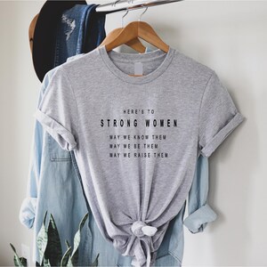 May include: Gray t-shirt with the text "HERE'S TO STRONG WOMEN MAY WE KNOW THEM MAY WE BE THEM MAY WE RAISE THEM" printed in black.