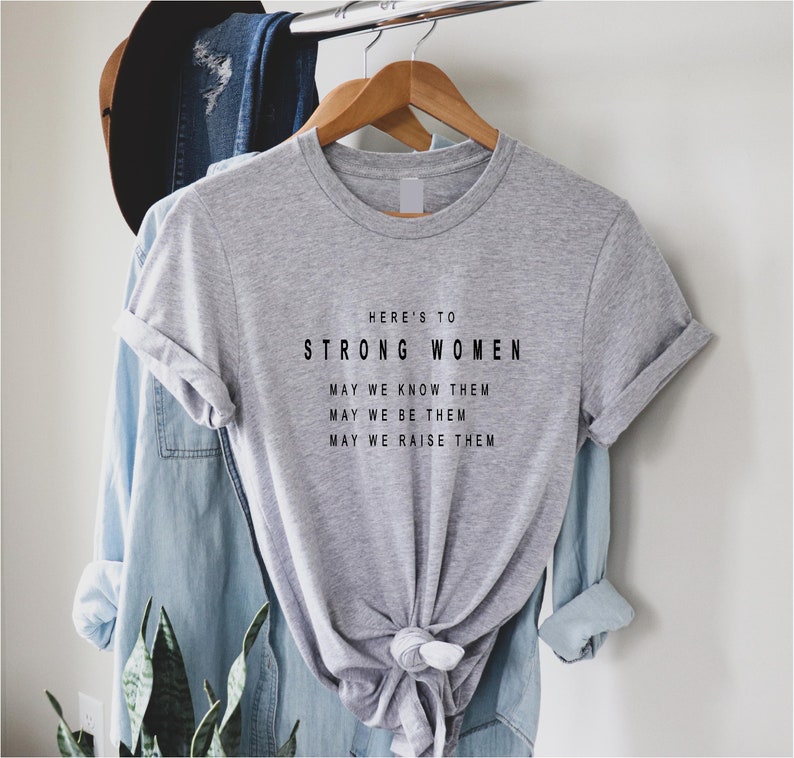 May include: Gray t-shirt with the text "HERE'S TO STRONG WOMEN MAY WE KNOW THEM MAY WE BE THEM MAY WE RAISE THEM" printed in black.
