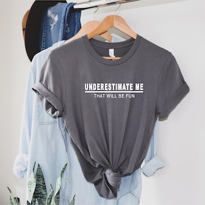 May include: A grey t-shirt with the text "UNDERESTIMATE ME THAT WILL BE FUN" printed in white.