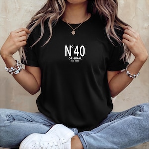 May include: Black t-shirt with the text "N&deg; 40 ORIGINAL EST 1986" in white. The shirt is worn by a person wearing blue jeans and white sneakers. The person is also wearing bracelets and a necklace.