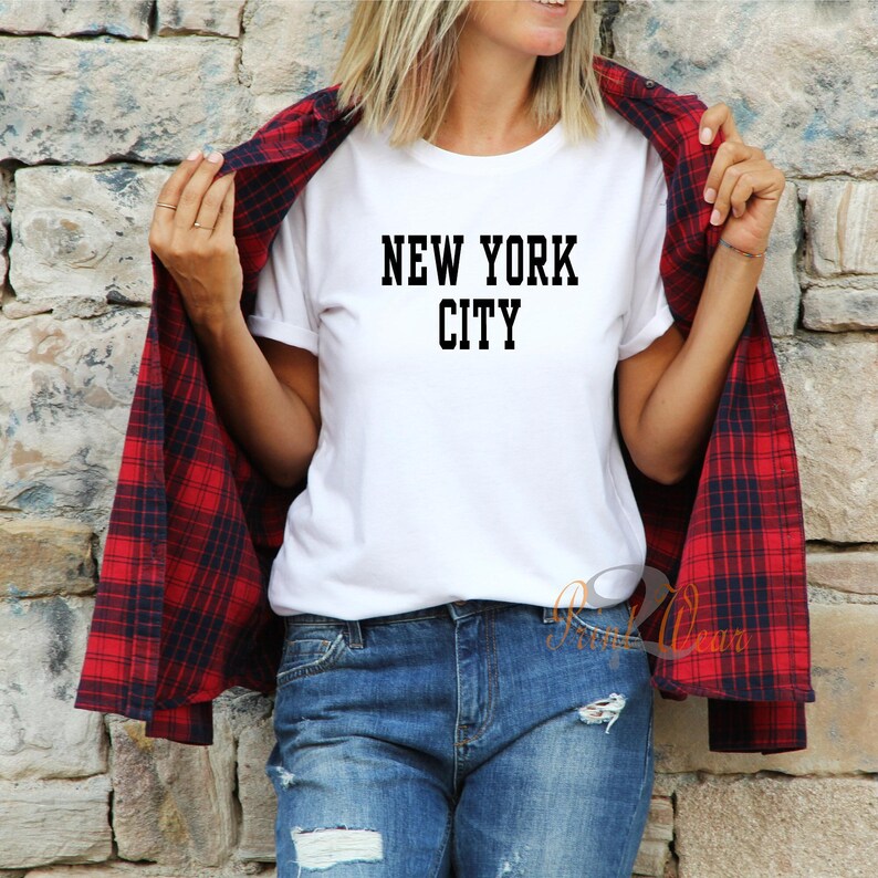 Puede incluir: Una camiseta blanca con texto negro que dice "NEW YORK CITY". La camiseta la lleva puesta una persona con una camisa de cuadros rojos y negros encima.