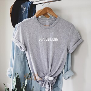 May include: A gray t-shirt with the text "Blah, Blah, Blah" printed on the front. The shirt is hanging on a wooden hanger.