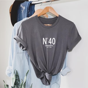 May include: A dark gray t-shirt with rolled-up sleeves, featuring the text "N&deg; 40 ORIGINAL EST 1986" in white. The shirt is hanging on a wooden hanger, with other clothing items visible in the background.