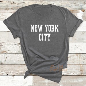 Puede incluir: Una camiseta gris oscura con el texto "NEW YORK CITY" impreso en blanco.