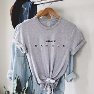 Może przedstawiać: Szary melanżowy t-shirt z kr&oacute;tkim rękawem i okrągłym dekoltem, ozdobiony czarnym napisem INHALE w g&oacute;rnym wierszu oraz E X H A L E w dolnym wierszu. Koszulka jest zawieszona na drewnianym wieszaku, a jej d&oacute;ł jest zawiązany w supeł z przodu.