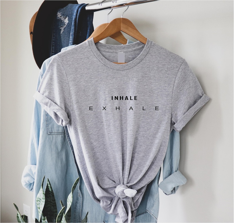 Może przedstawiać: Szary melanżowy t-shirt z kr&oacute;tkim rękawem i okrągłym dekoltem, ozdobiony czarnym napisem INHALE w g&oacute;rnym wierszu oraz E X H A L E w dolnym wierszu. Koszulka jest zawieszona na drewnianym wieszaku, a jej d&oacute;ł jest zawiązany w supeł z przodu.