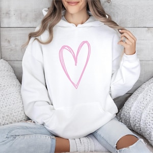 May include: White hoodie featuring a large, light pink heart design. The hoodie has a drawstring hood and a kangaroo pocket. The person is wearing light blue jeans.
