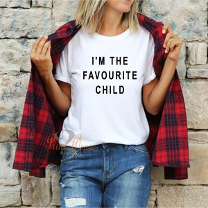 Peut inclure: T-shirt blanc avec un texte noir qui dit "Je suis l'enfant pr&eacute;f&eacute;r&eacute;". Une personne porte le t-shirt avec une chemise &agrave; carreaux rouge et noir par-dessus.