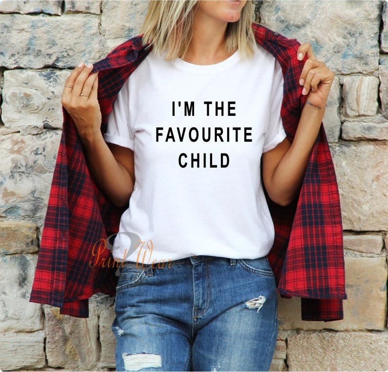 Peut inclure: T-shirt blanc avec un texte noir qui dit "Je suis l'enfant pr&eacute;f&eacute;r&eacute;". Une personne porte le t-shirt avec une chemise &agrave; carreaux rouge et noir par-dessus.
