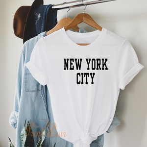 Puede incluir: Camiseta blanca con texto negro que dice "NEW YORK CITY". La camiseta est&aacute; colgada en una percha de madera.