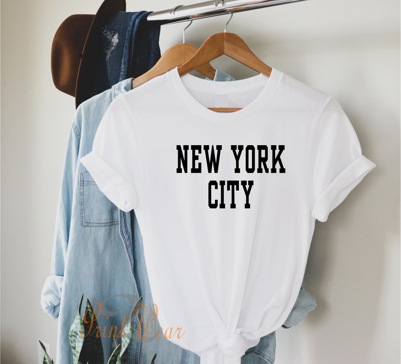 Puede incluir: Camiseta blanca con texto negro que dice "NEW YORK CITY". La camiseta est&aacute; colgada en una percha de madera.