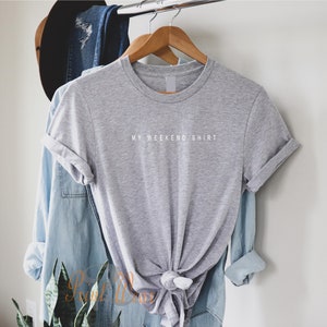 May include: A gray t-shirt with the text "MY WEEKEND SHIRT" printed in white on the front. The shirt is hanging on a wooden hanger.