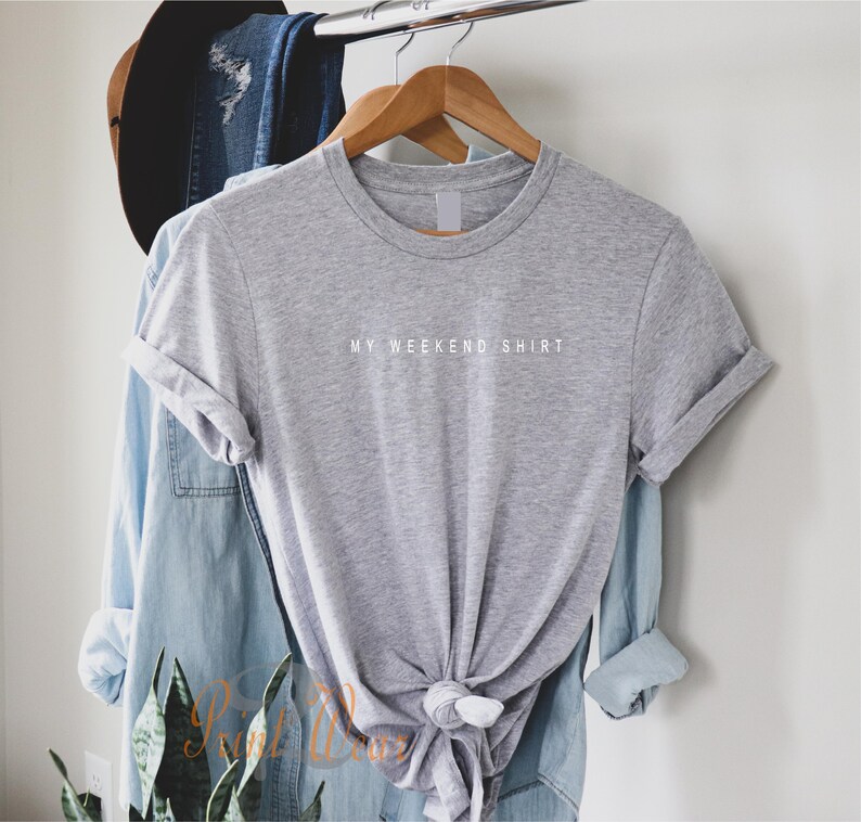 May include: A gray t-shirt with the text "MY WEEKEND SHIRT" printed in white on the front. The shirt is hanging on a wooden hanger.