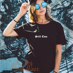 May include: A black t-shirt with the text "Still Emo" in white lettering. The shirt is being worn by a person with brown hair and blue sunglasses.