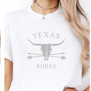May include: White t-shirt with a gray graphic design featuring a bull skull with crossed arrows and the text "TEXAS RODEO".