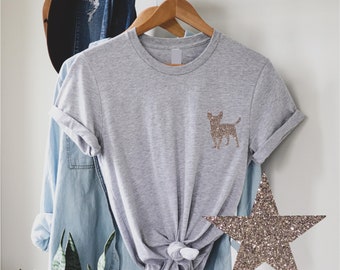 May include: A gray t-shirt with a gold glitter chihuahua silhouette on the front. The shirt is tied at the waist and is hanging on a hanger.