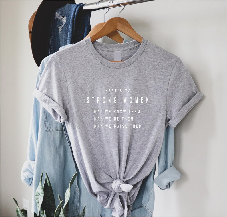 May include: A gray t-shirt with the text "HERE'S TO STRONG WOMEN MAY WE KNOW THEM MAY WE BE THEM MAY WE RAISE THEM" printed in white.