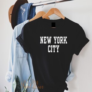 Puede incluir: Camiseta negra con texto blanco que dice "NEW YORK CITY" colgada de una percha de madera.