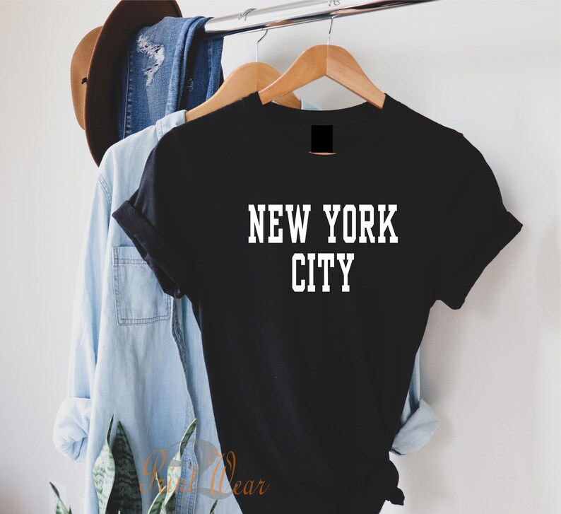 Puede incluir: Camiseta negra con texto blanco que dice "NEW YORK CITY" colgada de una percha de madera.