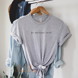 May include: A gray t-shirt with the text "MY WEEKEND SHIRT" printed on the front. The shirt is hanging on a wooden hanger.