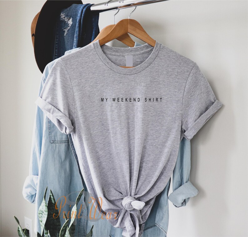 May include: A gray t-shirt with the text "MY WEEKEND SHIRT" printed on the front. The shirt is hanging on a wooden hanger.