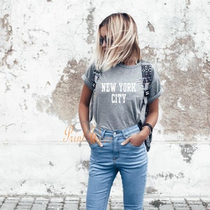 Puede incluir: Una camiseta gris con el texto "NEW YORK CITY" en letras blancas. La camiseta la lleva puesta una persona con el pelo rubio y unos vaqueros azules.