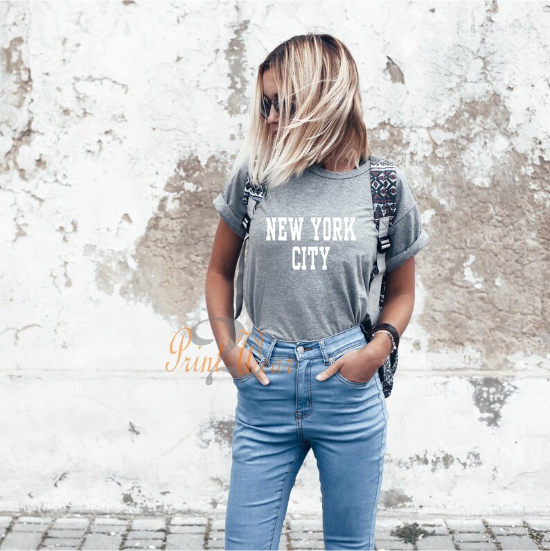 Puede incluir: Una camiseta gris con el texto "NEW YORK CITY" en letras blancas. La camiseta la lleva puesta una persona con el pelo rubio y unos vaqueros azules.