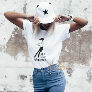 May include: White t-shirt featuring a black ska graphic and the text "SKA THE ORIGINAL RUDE GIRL", with a checkered pattern. The model is wearing a white baseball cap with black star designs and blue jeans.