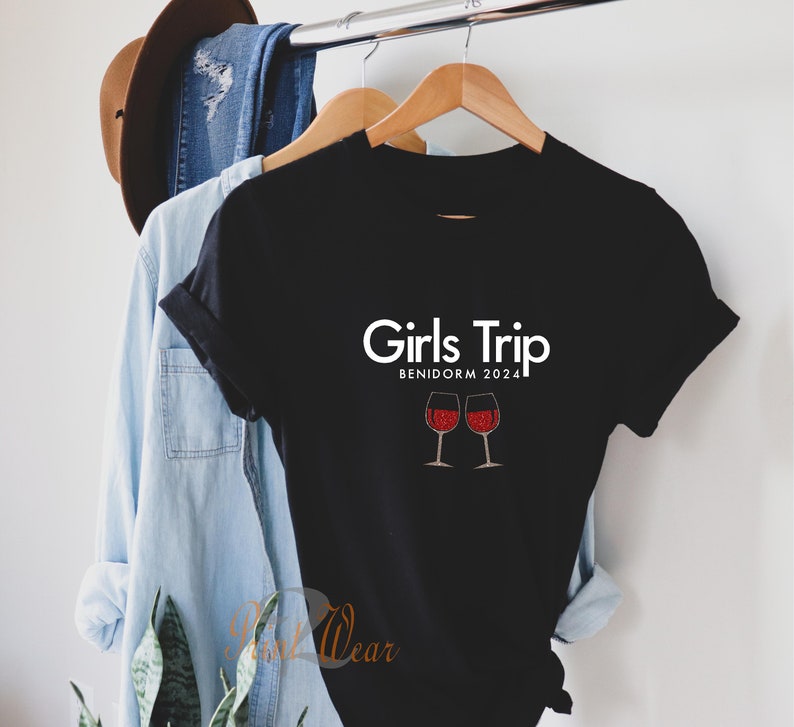 Pode incluir: Camiseta preta com texto branco que diz "Girls Trip Benidorm 2024" e duas ta&ccedil;as de vinho tinto com glitter.