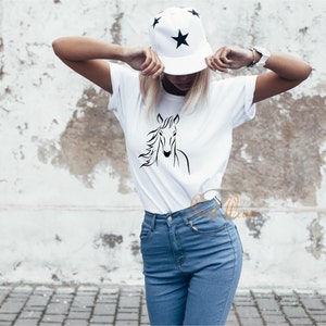 K&ouml;nnte beinhalten: Ein wei&szlig;es T-Shirt mit einer schwarzen Linienzeichnung eines Pferdekopfes auf der Vorderseite. Das Model tr&auml;gt eine blaue Jeans und eine schwarz-wei&szlig;e Baseballkappe mit Sternenmuster.