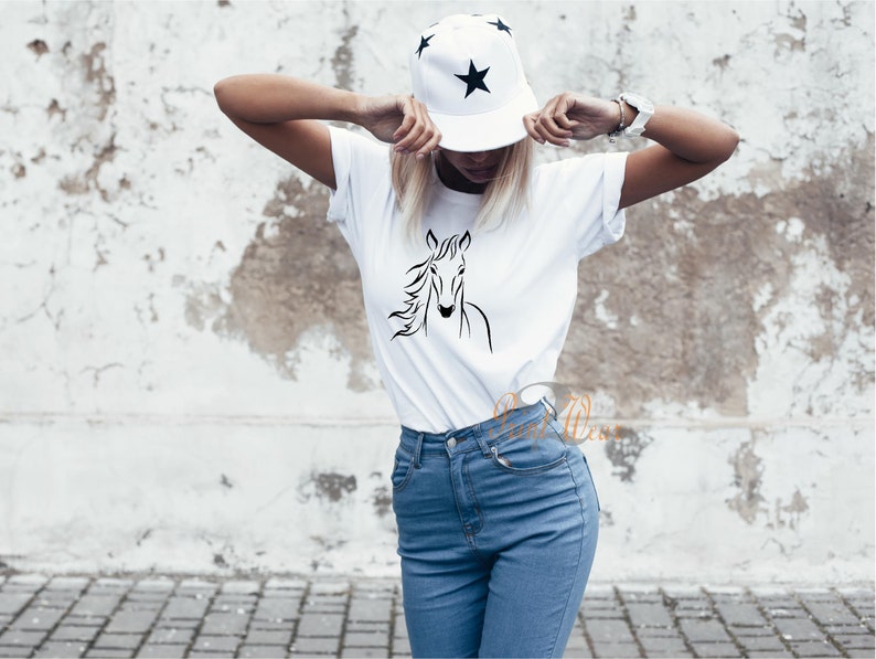 K&ouml;nnte beinhalten: Ein wei&szlig;es T-Shirt mit einer schwarzen Linienzeichnung eines Pferdekopfes auf der Vorderseite. Das Model tr&auml;gt eine blaue Jeans und eine schwarz-wei&szlig;e Baseballkappe mit Sternenmuster.