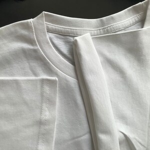 Może przedstawiać: Gładki biały t-shirt z klasycznym okrągłym dekoltem, prążkowanym kołnierzykiem i kr&oacute;tkimi rękawami. Odzież wykonana jest z gładkiej, jednolicie białej tkaniny z widocznymi detalami podw&oacute;jnych szw&oacute;w na krawędziach rękaw&oacute;w i przy dekolcie.