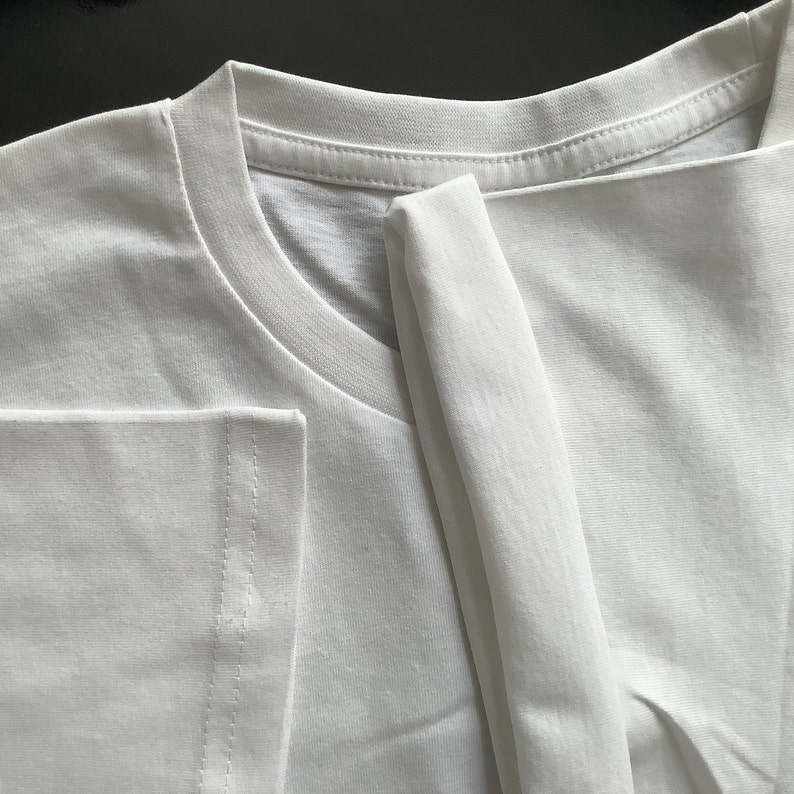 Może przedstawiać: Gładki biały t-shirt z klasycznym okrągłym dekoltem, prążkowanym kołnierzykiem i kr&oacute;tkimi rękawami. Odzież wykonana jest z gładkiej, jednolicie białej tkaniny z widocznymi detalami podw&oacute;jnych szw&oacute;w na krawędziach rękaw&oacute;w i przy dekolcie.