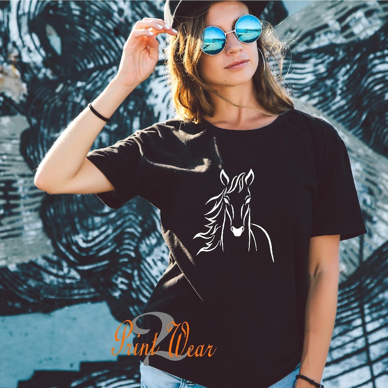 K&ouml;nnte beinhalten: Ein schwarzes T-Shirt mit einem wei&szlig;en Umriss eines Pferdekopfes auf der Vorderseite. Das Model tr&auml;gt das Shirt und eine blaue Sonnenbrille.
