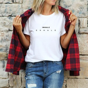 Może przedstawiać: Biały t-shirt z kr&oacute;tkim rękawem z czarnym napisem INHALE EXHALE na piersi, noszony pod koszulą z długim rękawem w czerwono-czarną kratę oraz niebieskimi dżinsami z przetarciami i dziurami z przodu.