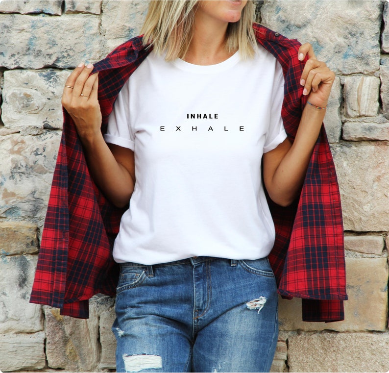 Może przedstawiać: Biały t-shirt z kr&oacute;tkim rękawem z czarnym napisem INHALE EXHALE na piersi, noszony pod koszulą z długim rękawem w czerwono-czarną kratę oraz niebieskimi dżinsami z przetarciami i dziurami z przodu.