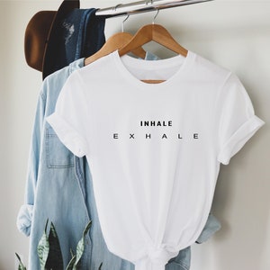 Może przedstawiać: Biały t-shirt z kr&oacute;tkim rękawem, okrągłym dekoltem i podwiniętymi rękawami. Z przodu koszulki znajduje się czarny napis INHALE wyśrodkowany nad tekstem E X H A L E, w kt&oacute;rym poszczeg&oacute;lne litery są oddzielone od siebie spacjami.