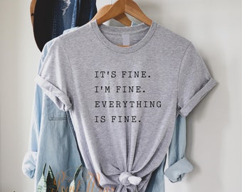 May include: A heather gray short-sleeved t-shirt with a crew neckline. The shirt features black text printed on the front that reads: IT'S FINE. I'M FINE. EVERYTHING IS FINE. The hem of the shirt is tied in a knot at the center front.