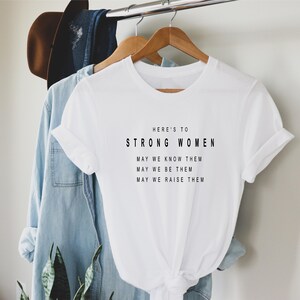 May include: White t-shirt with black text that reads "Here's to Strong Women. May we know them. May we be them. May we raise them."